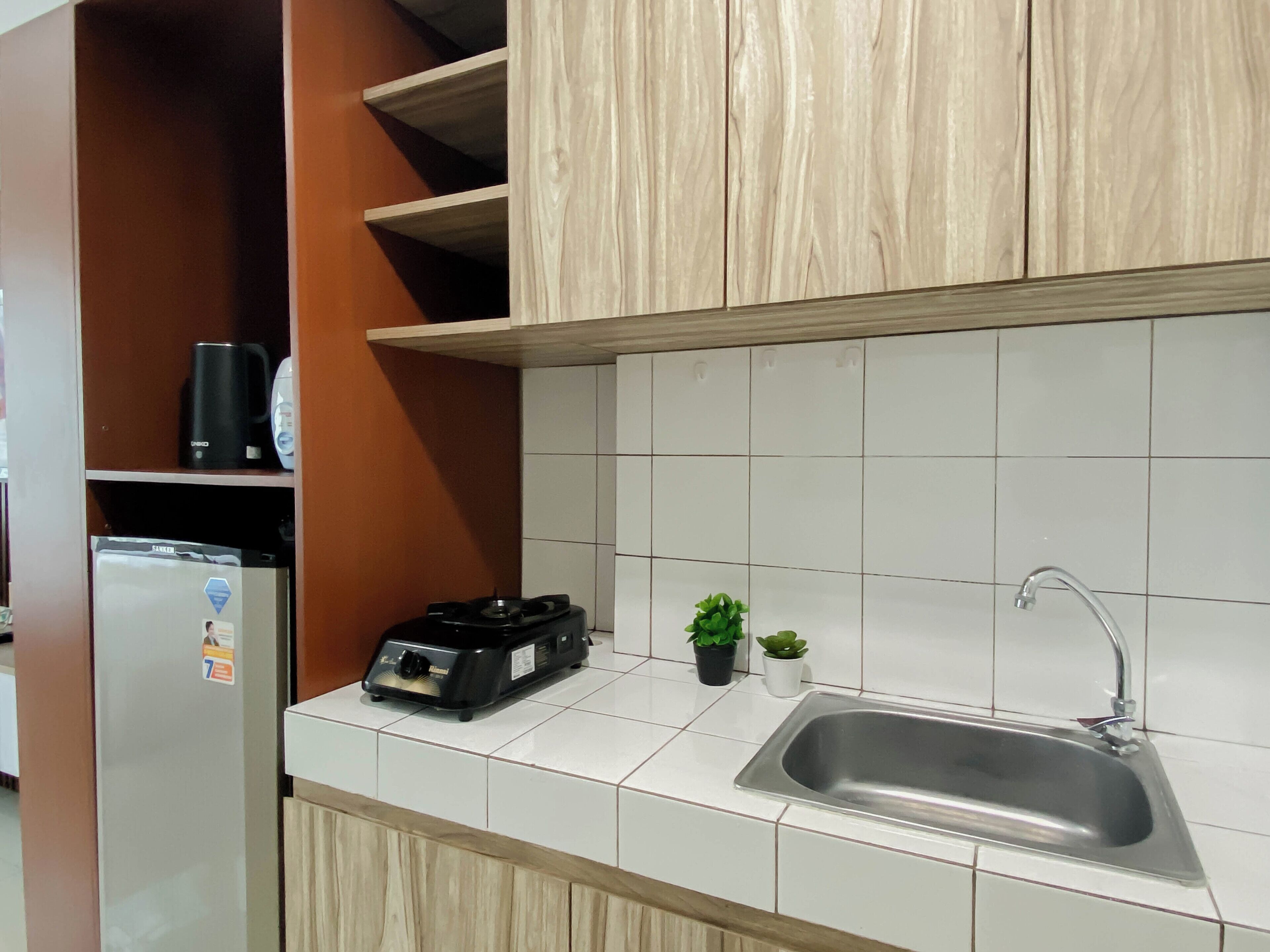 Cocina con baldosas blancas, un lavamanos de acero inoxidable, armarios de madera, una cafetera negra y una pequeña planta en maceta.