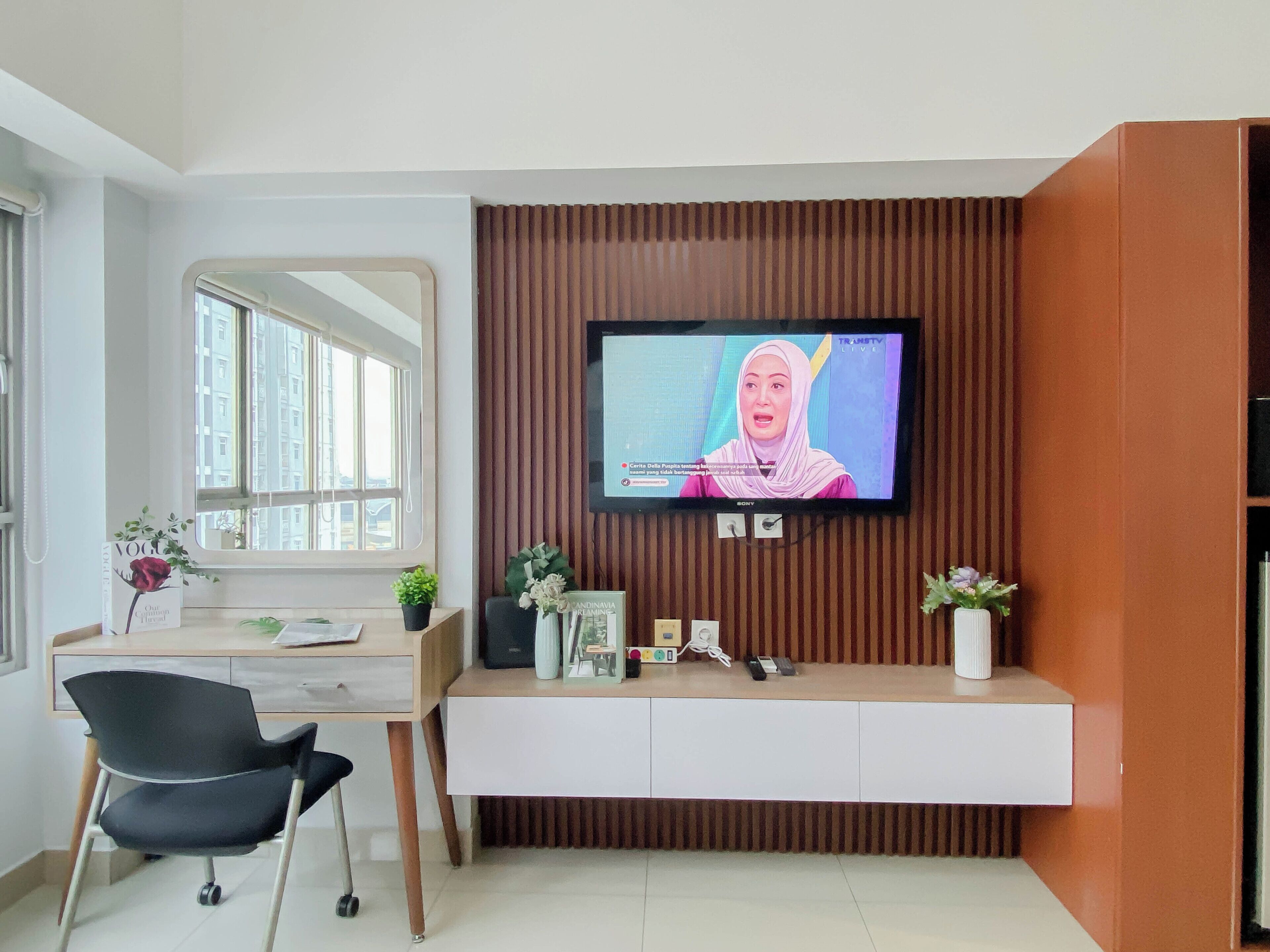 A modern living room with a TV mounted on a wooden panel wall, a desk with a chair, and a window with a view of buildings.