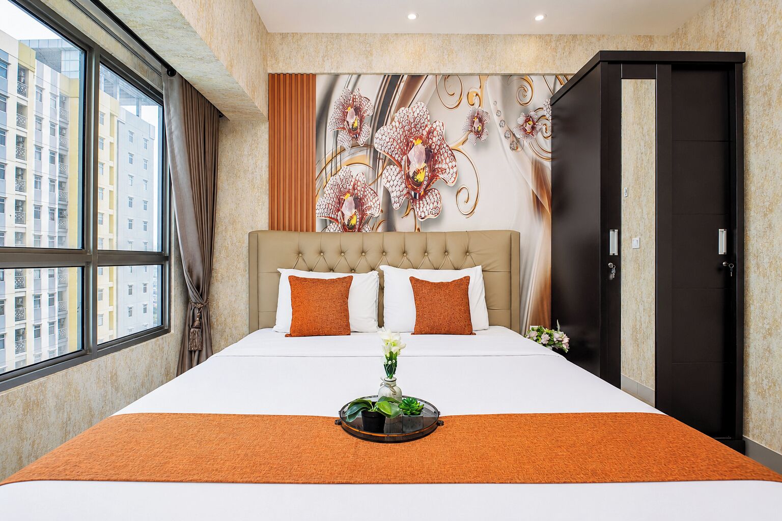 A hotel room with a large bed, two pillows, a nightstand with a plant, a wall mural, and a view of buildings outside.