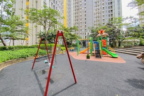 A playground with swings and a slide, surrounded by greenery and tall buildings. - Calming And Comfortable Living In 3Br At Apartment Springlake Summarecon Bekasi (Bekasi)