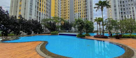 A swimming pool with a wooden deck, surrounded by greenery and tall residential buildings.