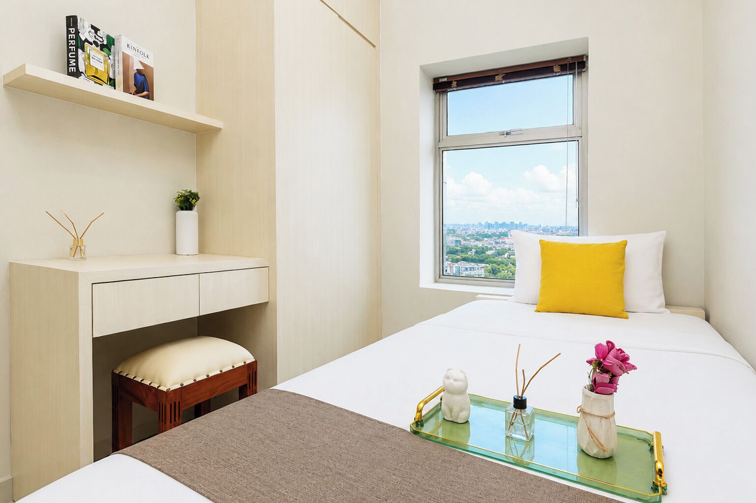 A modern hotel room with a bed, a window with a city view, a small desk with a shelf, and a bedside table with a tray.