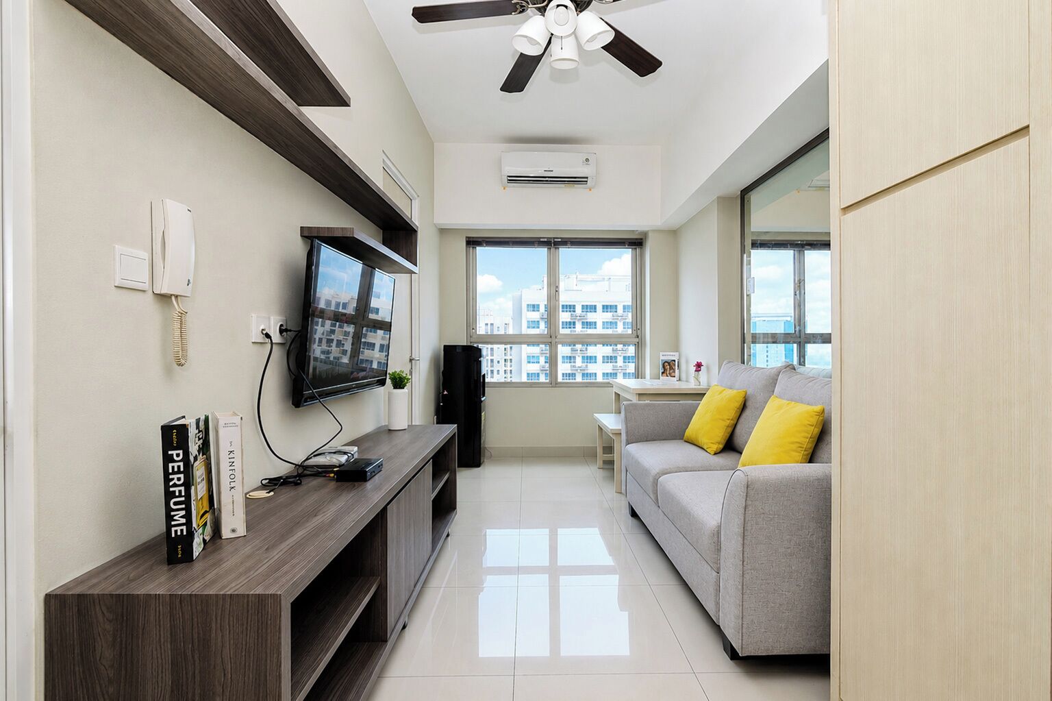 Una sala moderna con televisor de pantalla plana, mueble de madera para entretenimiento, sofá gris con cojines amarillos, ventilador de techo y ventanas amplias con vistas a la ciudad.