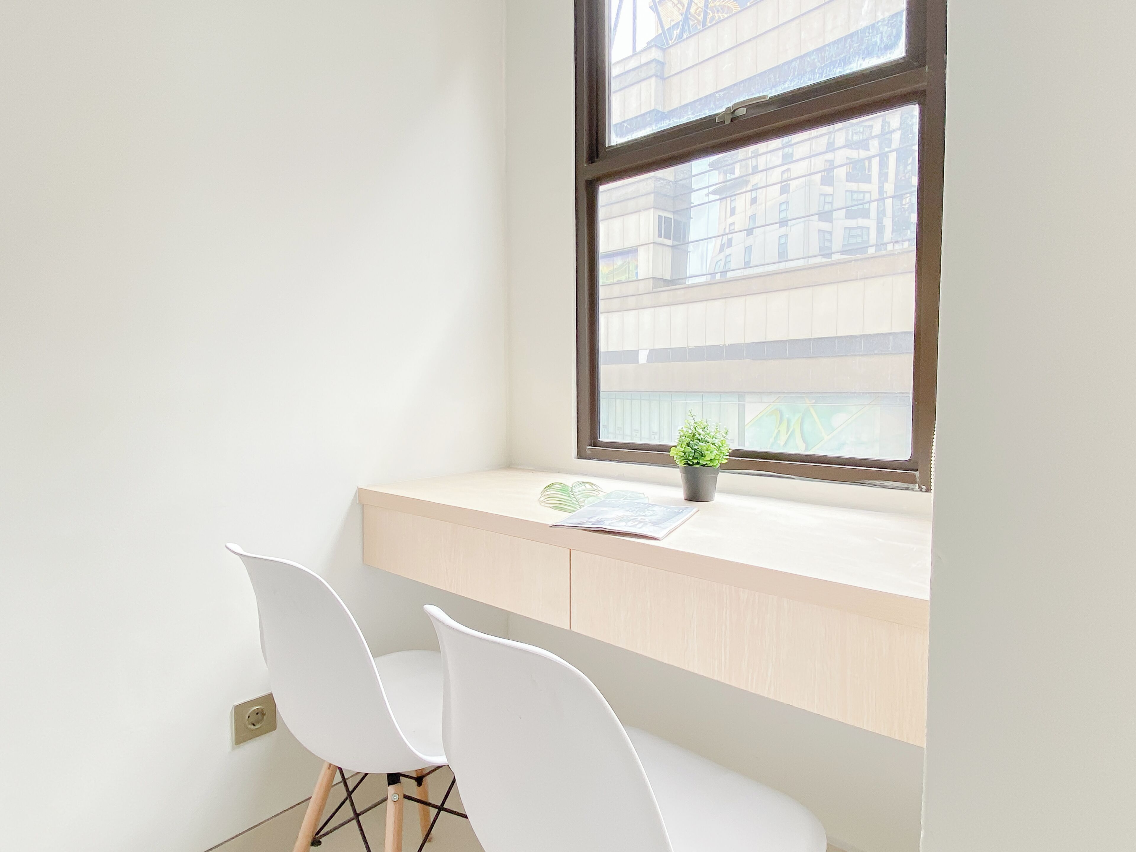 A corner desk with two white chairs, a small potted plant, and a window with a view of buildings.