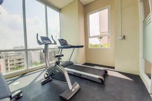 A gym with a treadmill and an elliptical machine, large windows with a view of buildings, and a small window with a plant. - Splendid Studio At Apartment B Residence (Tangerang)