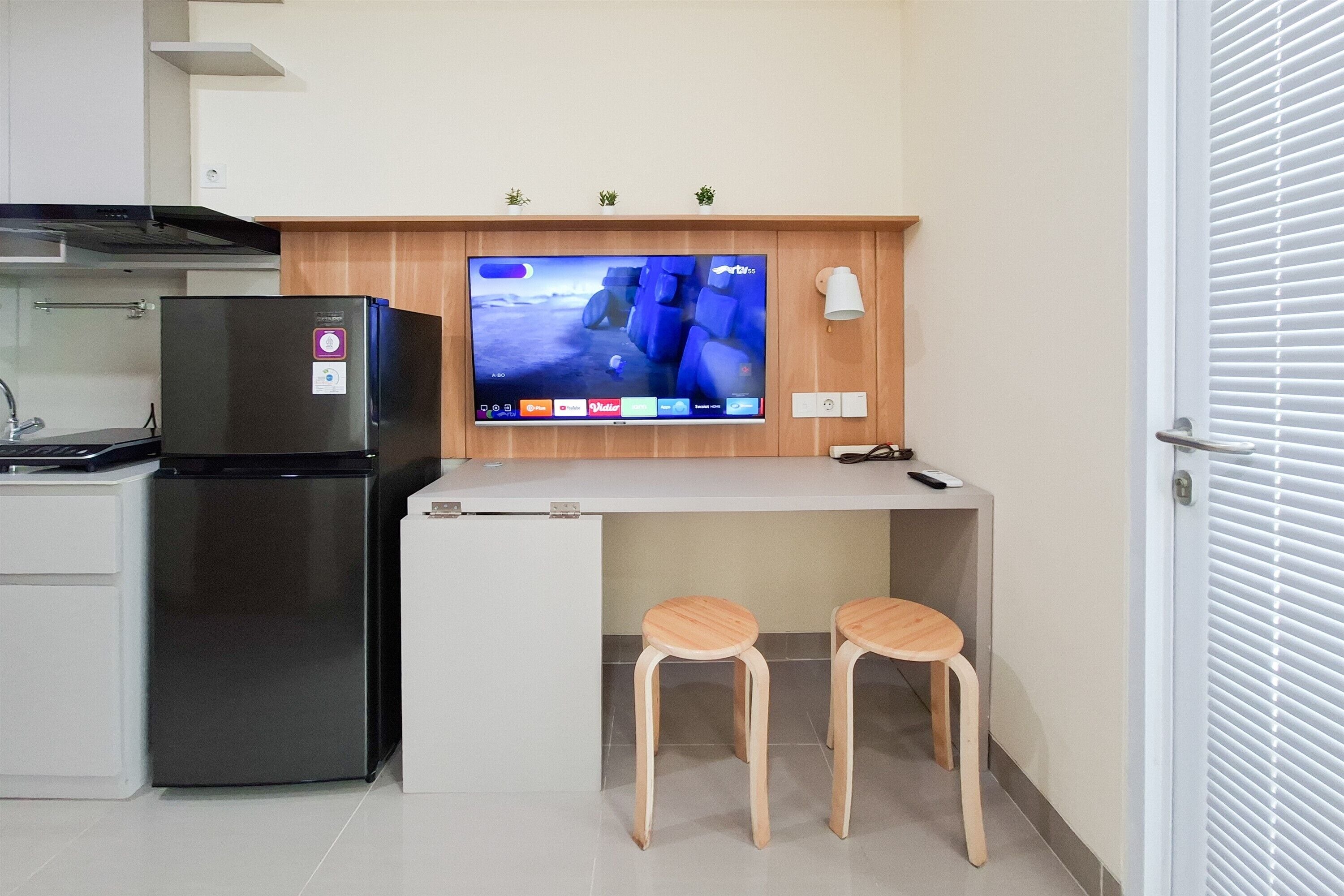 Una kitchenette compacta con refrigerador, un televisor de pantalla plana sobre una encimera blanca y dos taburetes de madera.