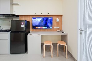A compact kitchenette with a refrigerator, a flat-screen TV mounted above a white countertop, and two wooden stools. - Comfortable And Homey Studio Room At Apartment B Residence (Tangerang)