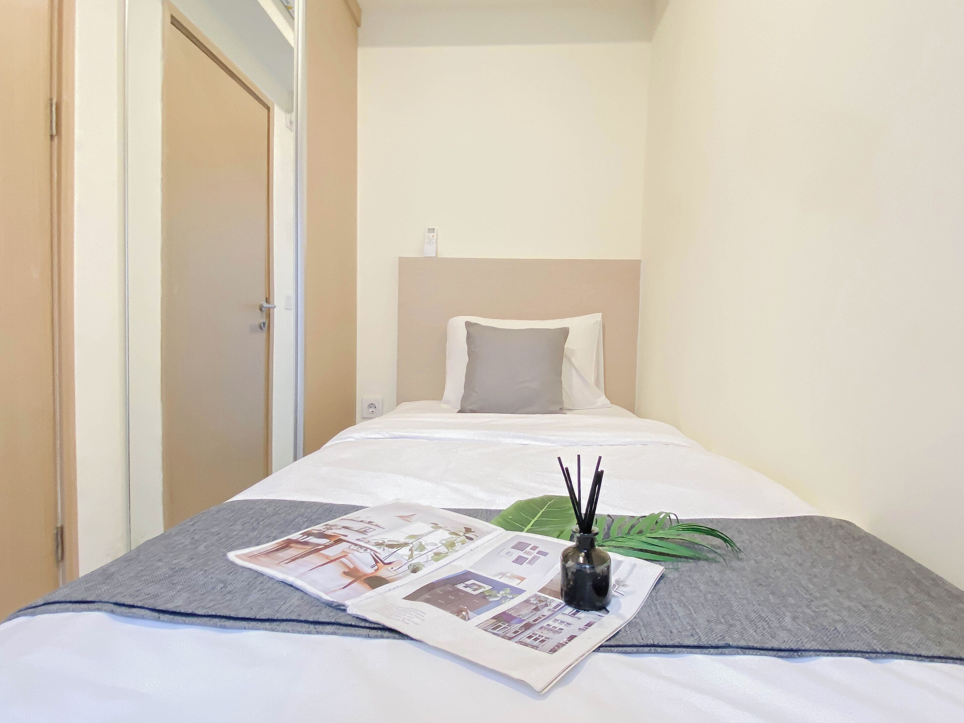 A hotel room with a bed, a bedside table with a magazine, a small vase with reeds, and a green plant.