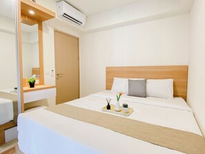 A hotel room with a bed, a bedside table, a mirror, an air conditioning unit, and a tray with decorative items. - Cozy Earth Tone 2Br At Apartment Meikarta District 1 (Cikarang)