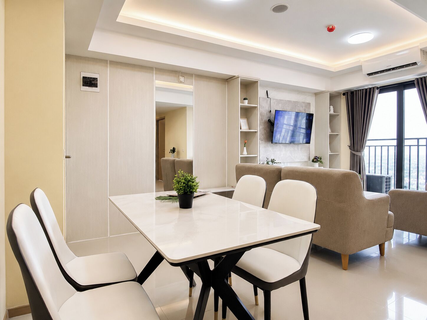 A modern living room with a dining area, a flat-screen TV mounted on the wall, and a balcony with curtains.