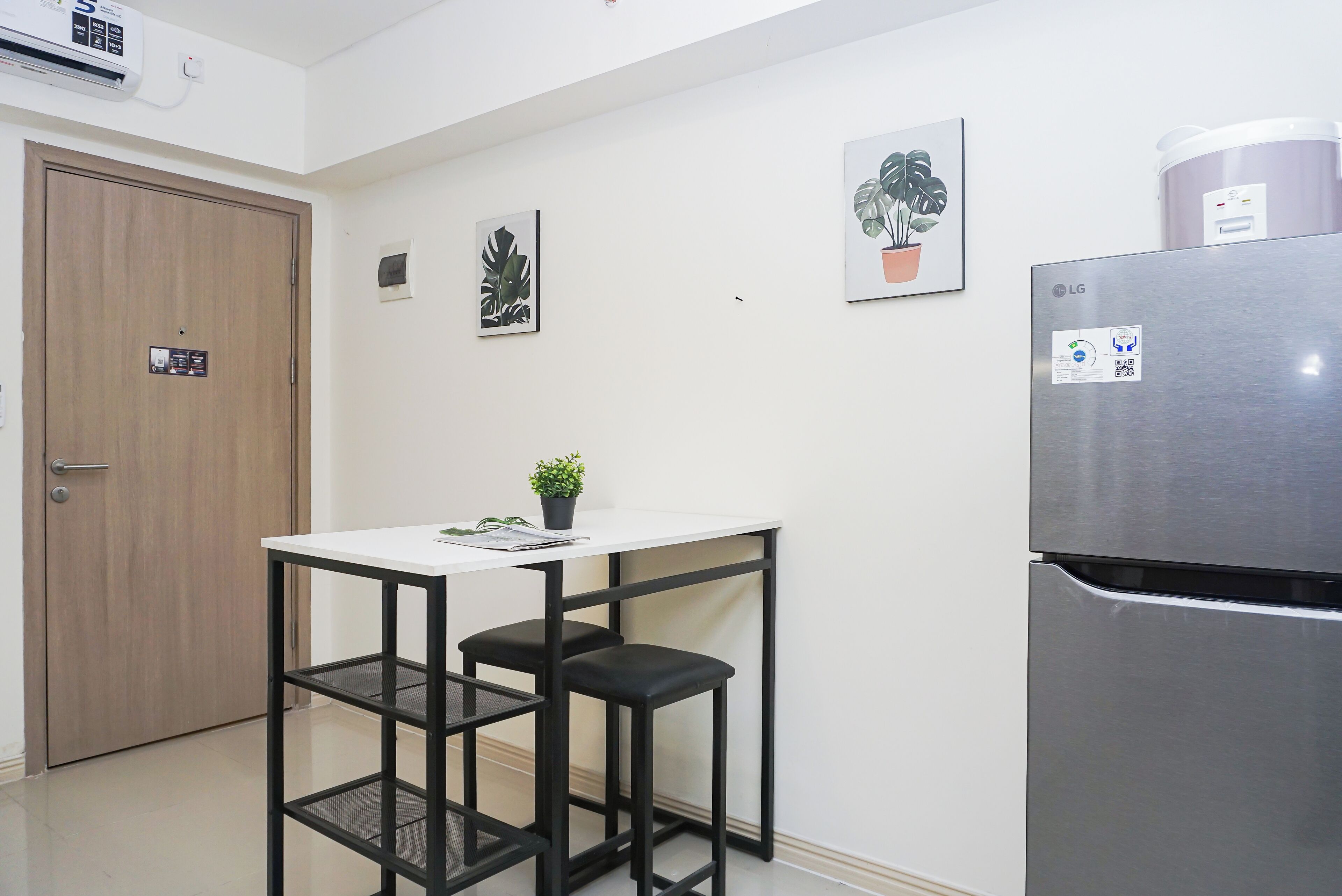 Cocina compacta con una mesa blanca, dos taburetes negros, un refrigerador de acero inoxidable y una obra de arte en la pared.