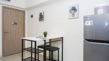 Cocina compacta con una mesa blanca, dos taburetes negros, un refrigerador de acero inoxidable y una obra de arte en la pared.