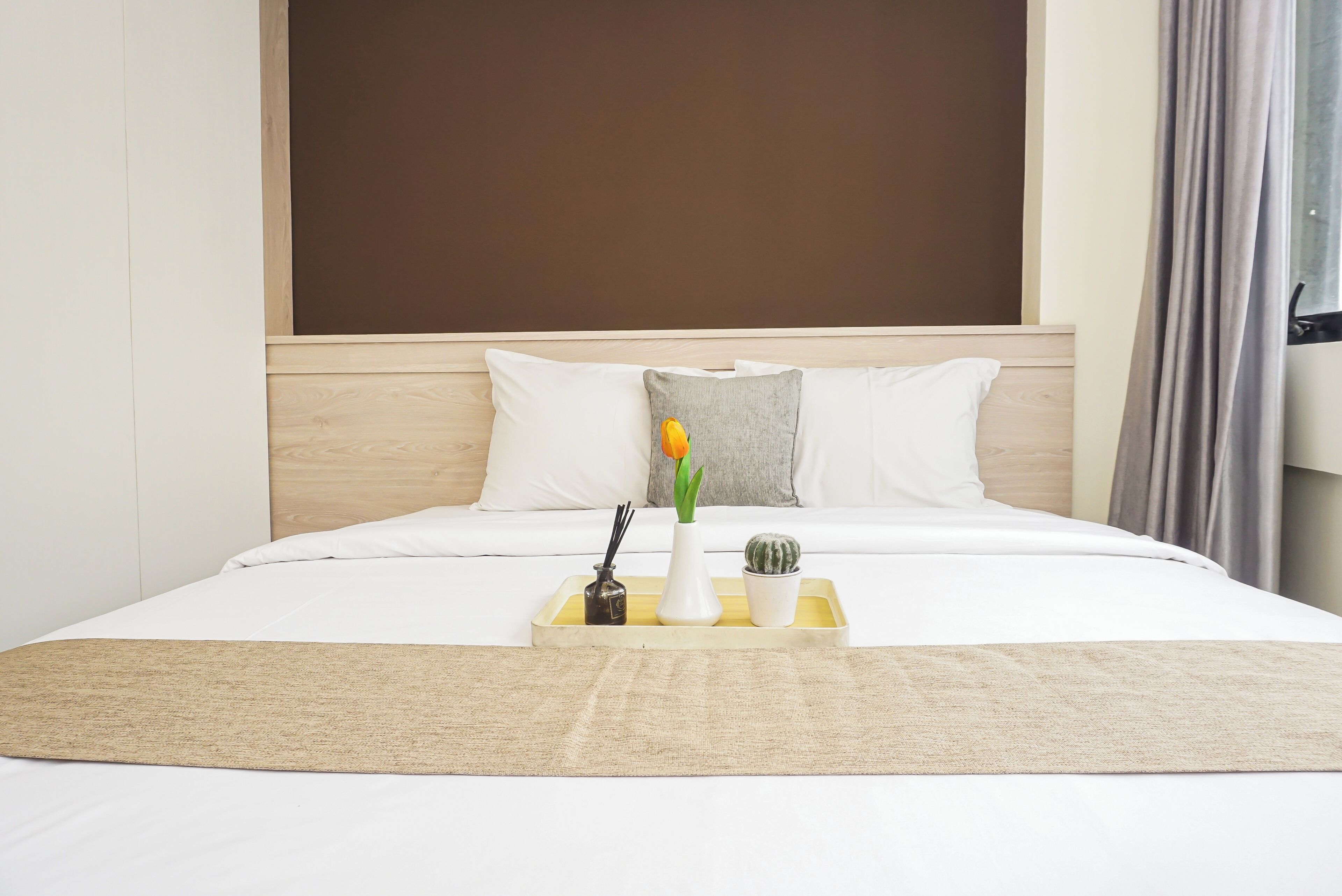Una habitación de hotel con cama, una mesita de noche con un jarrón de flores, una bandeja con una vela y una pequeña planta, y un cabecero.