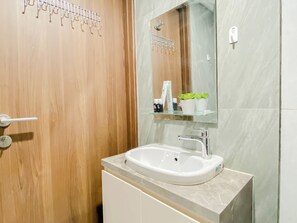 Une salle de bain avec une porte en bois, un lavabo blanc, un miroir et une étagère en verre où sont rangés les articles de toilette.