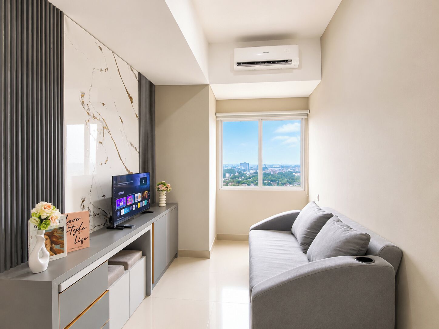 Un salon moderne avec un canapé gris, un téléviseur à écran plat posé sur une console grise, une vue sur la ville à travers une grande fenêtre et un appareil de climatisation fixé au plafond.