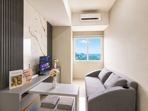 A modern living room with a grey sofa, a flat-screen TV, a white coffee table with a vase of flowers, and a city view through a large window. - Modern Style 1Br Apartment At Royal Sentul Park (Bogor)