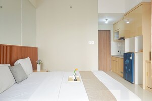 Habitación de hotel con cama, mesita de noche, un jarrón con flores, una mesita con una vela y una kitchenette.