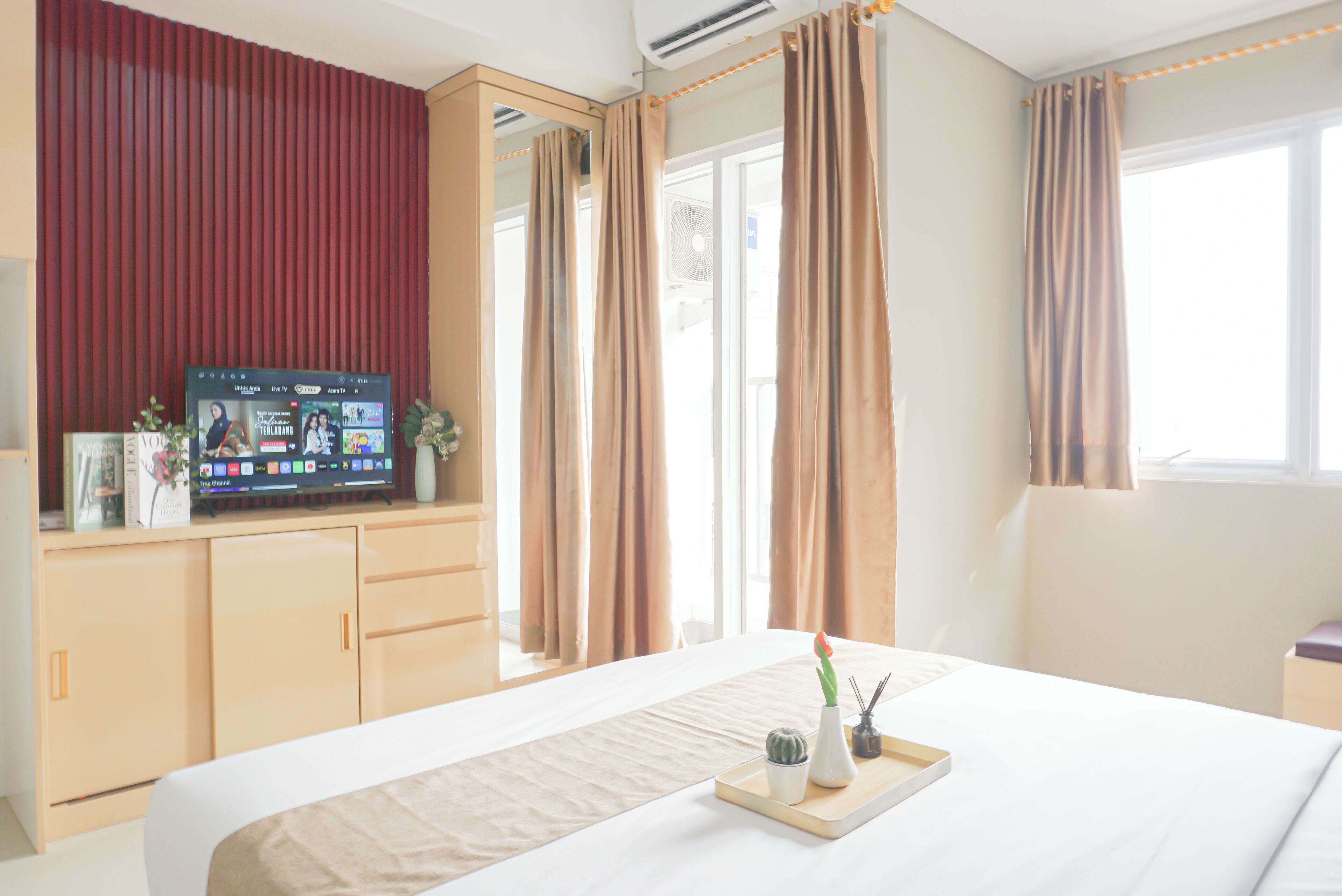 Una habitación de hotel con una cama, un televisor sobre un soporte, una ventana con cortinas y una mesita de noche con una bandeja.