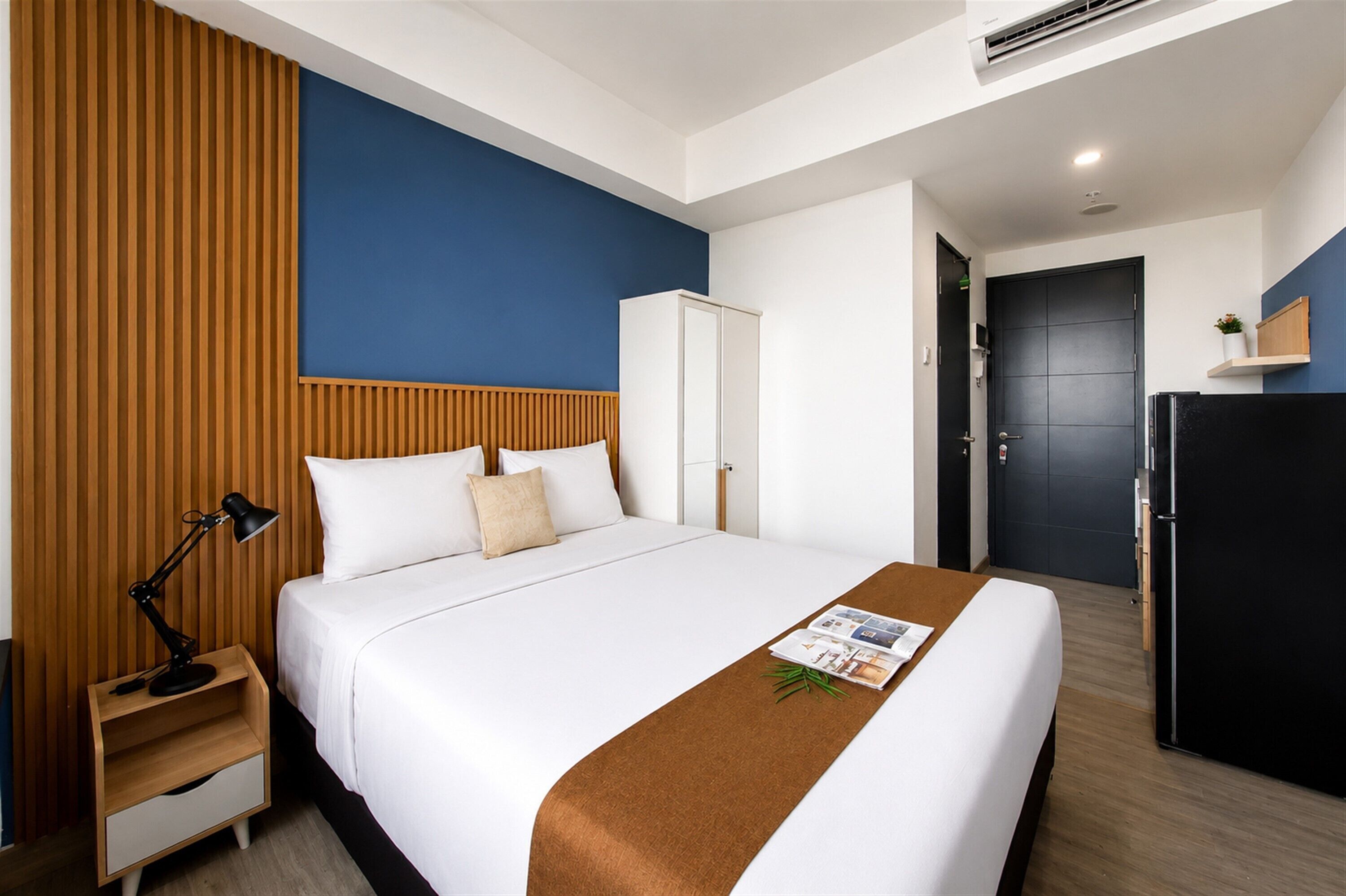 Een hotelkamer met een bed, nachtkastje, bureau, kledingkast, een zwarte koelkast en een blauwe wand.