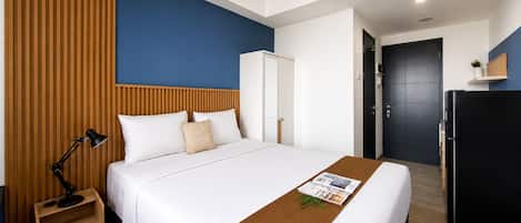 Une chambre d’hôtel avec un lit, une table de chevet, un bureau, une armoire, un réfrigérateur noir et un mur bleu.