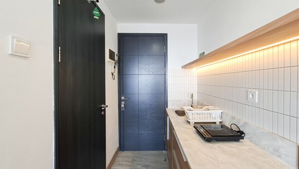A narrow hallway with a dark door, a white tiled wall, and a countertop with a basket and a small appliance. - Modern Cozy Studio At Apartment Collins Boulevard (Tangerang)