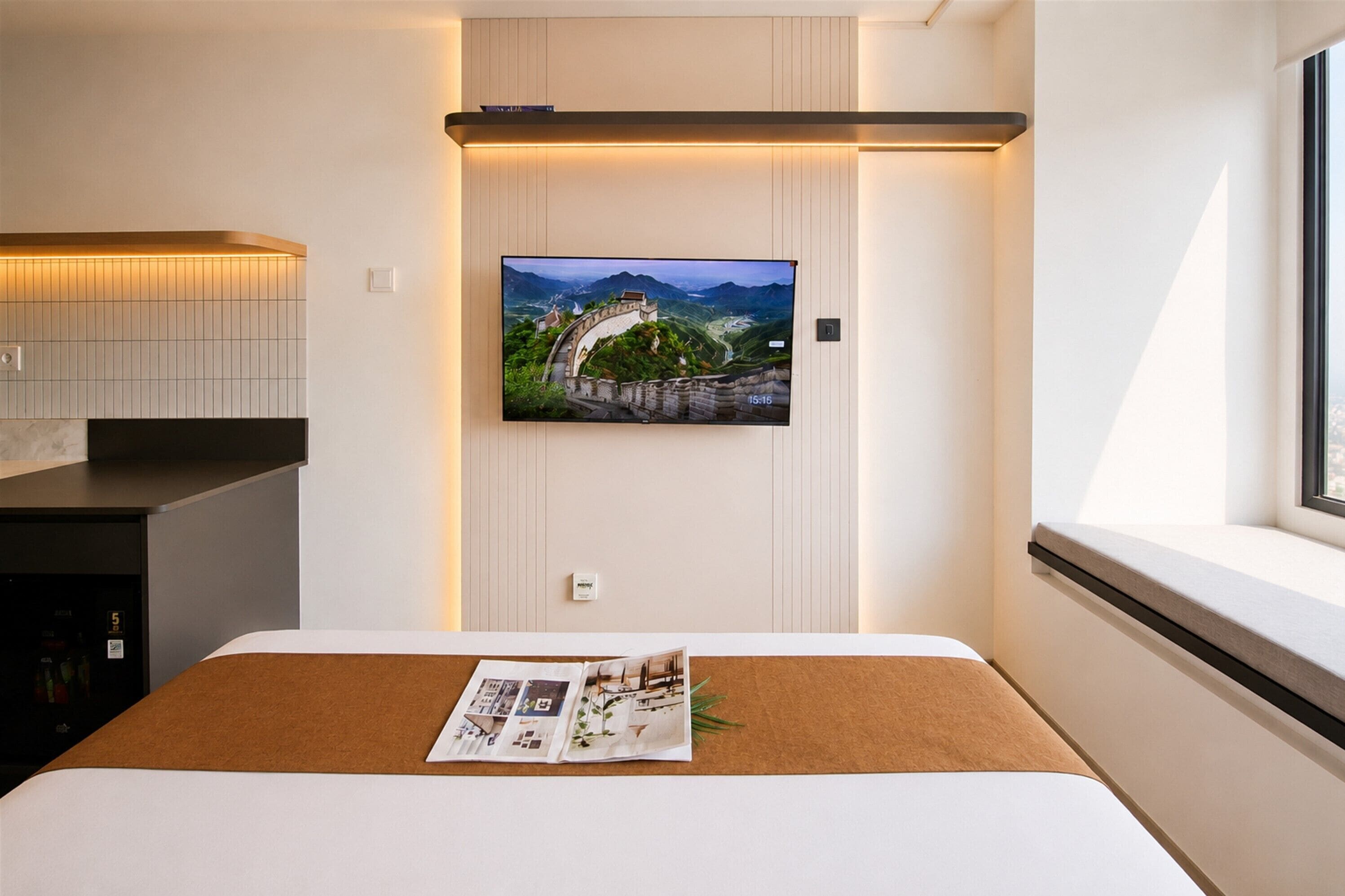 Quarto de hotel moderno com uma cama, televisão de tela plana na parede, janela com vista e uma pequena mesa com uma revista.