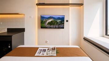 Quarto de hotel moderno com uma cama, televisão de tela plana na parede, janela com vista e uma pequena mesa com uma revista.