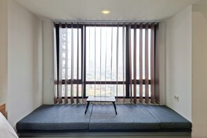 A room with a bed, a small table, and a view through large windows with blinds. - Great Deal Studio At Apartment Collins Boulevard (Tangerang)