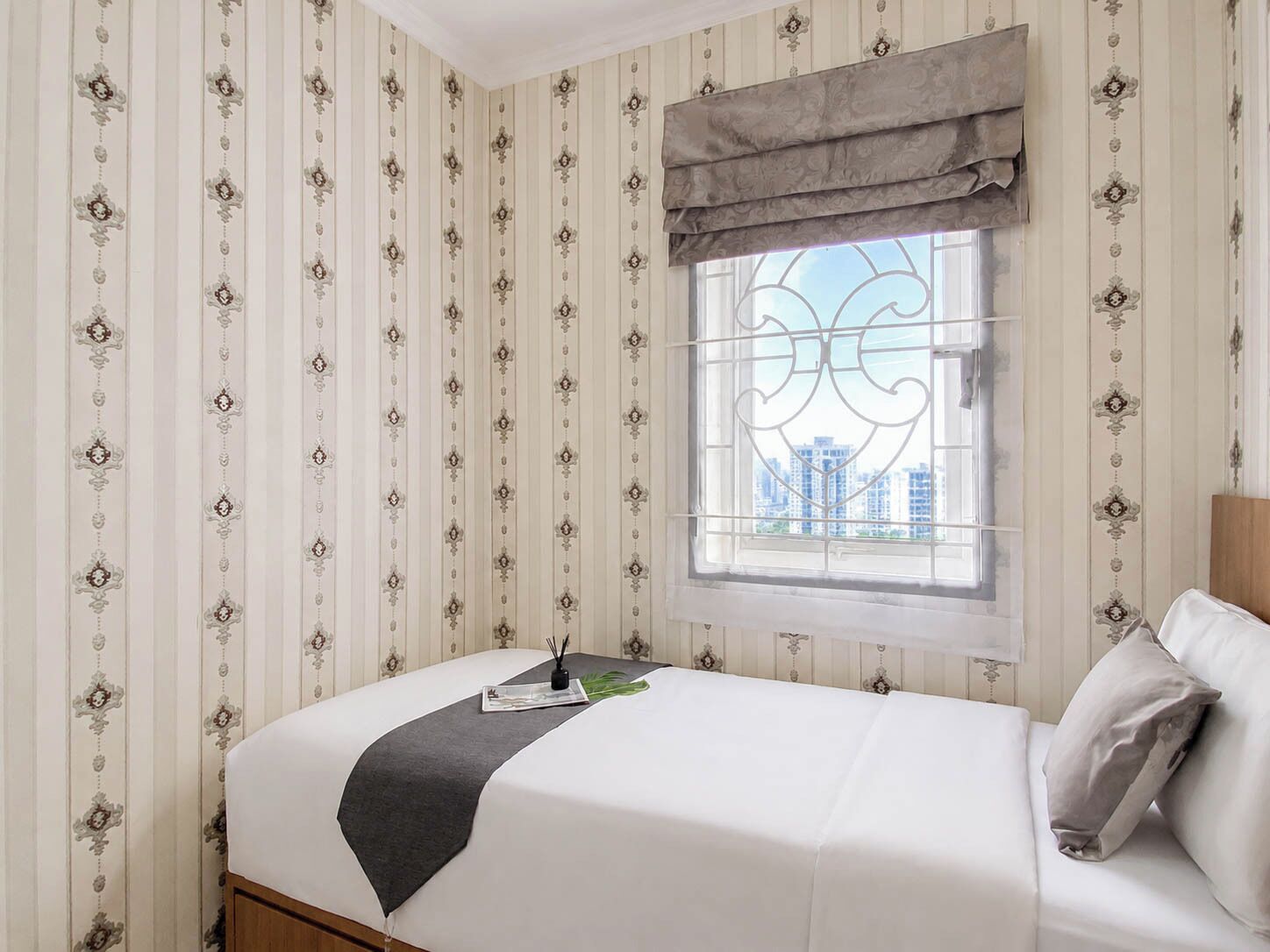 A neatly made bed with a gray throw, a window with a decorative valance, and striped wallpaper with a floral pattern.