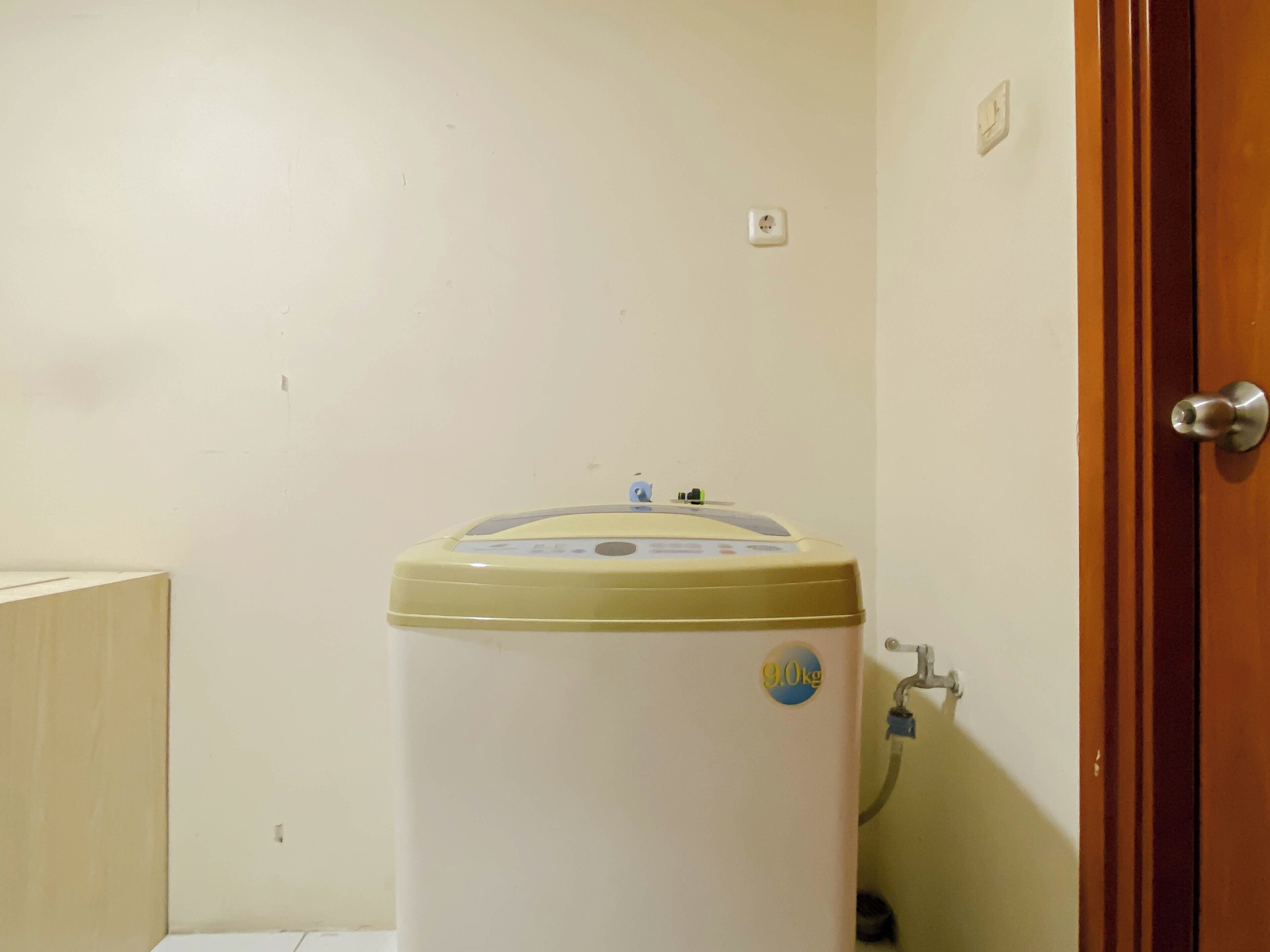 A compact, white water heater with a blue sticker indicating a weight of 9.0 kg, situated in a corner of a room with a wooden door and a white wall.