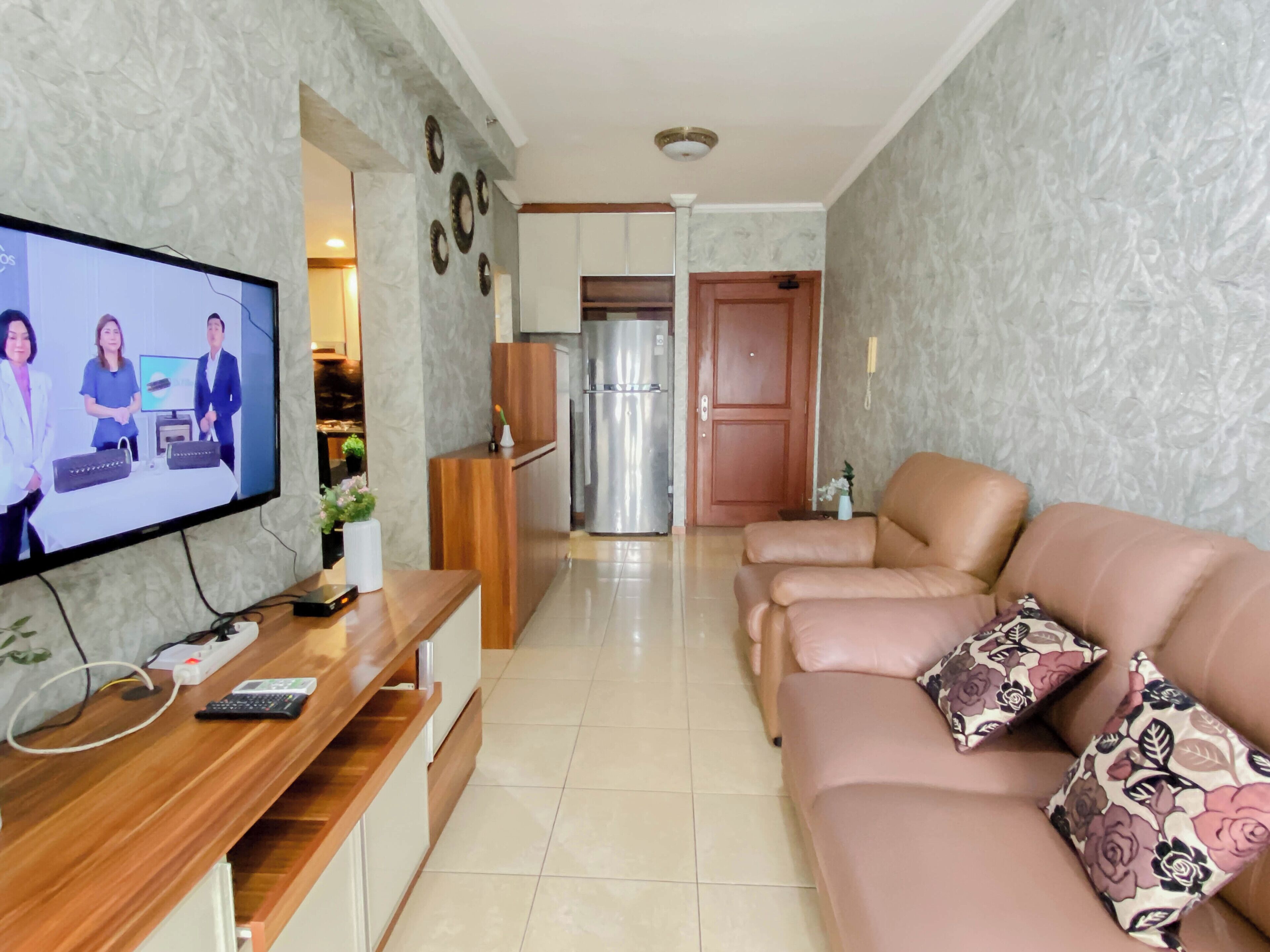 A living room with a television, wooden reception desk, leather sofa, and a kitchen in the background.