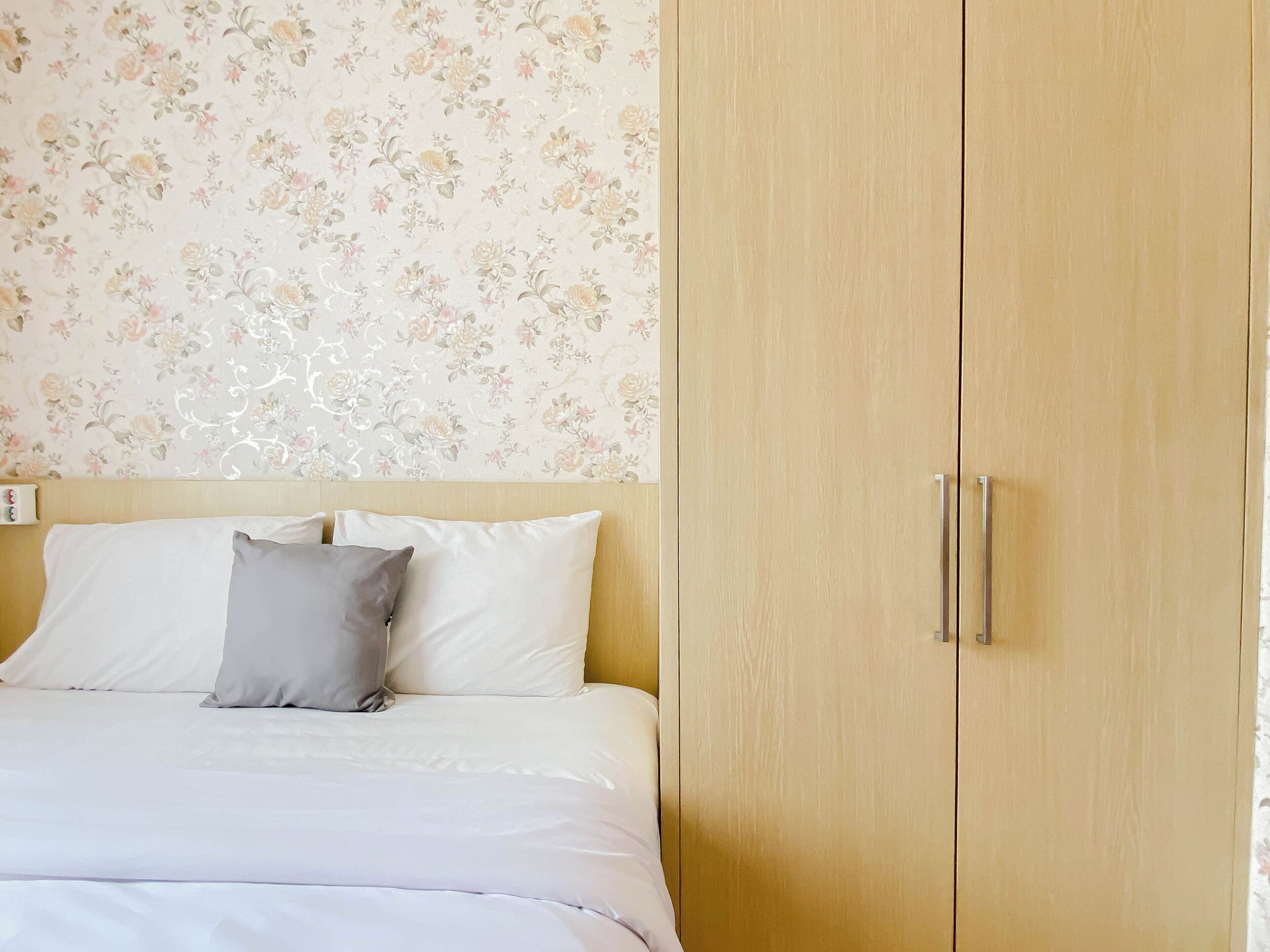 A bed with white bedding and a grey pillow, a wooden wardrobe with two doors, and a floral wallpaper pattern on the wall.