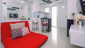 A modern living room with a red sofa, a dining area with a table and chairs, a TV mounted on the wall, and a large window. - Homey 2Br With Modern Interior At Apartment Bassura City (Jakarta)