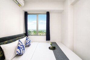 A hotel room with a bed, a window with curtains, a small table with a plant, and a wall-mounted air conditioner. - Homey 2Br With Modern Interior At Apartment Bassura City (Jakarta)