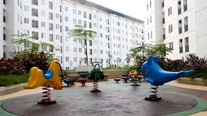 A playground with swings and a slide, surrounded by a building with multiple floors and balconies. - Homey 2Br With Modern Interior At Apartment Bassura City (Jakarta)