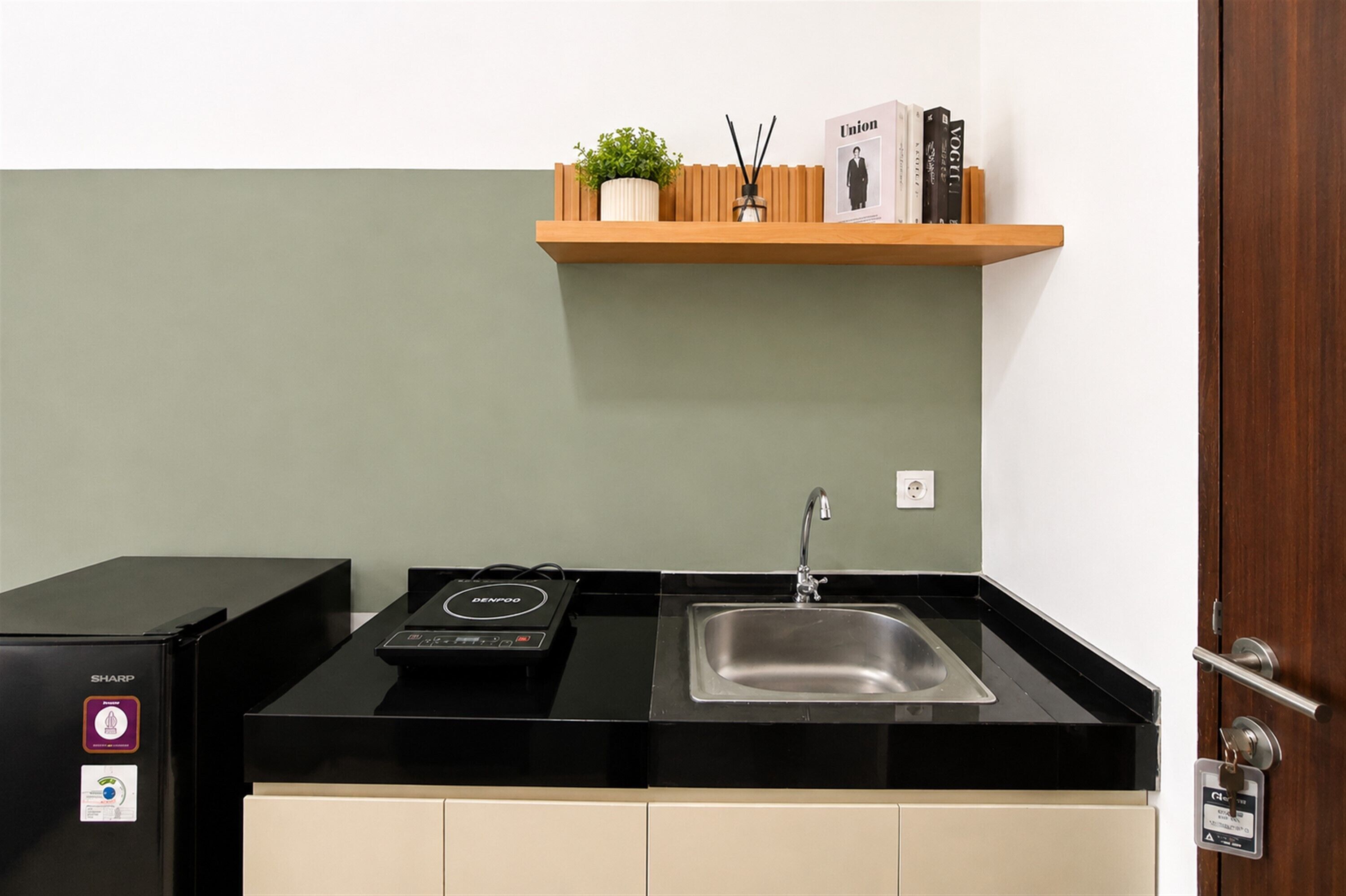 A compact kitchen with a black countertop, a stainless steel sink, a black refrigerator, and a wooden shelf with a plant and books.