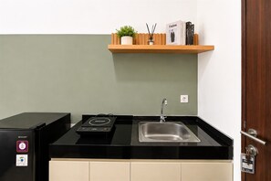 A compact kitchen with a black countertop, a stainless steel sink, a black refrigerator, and a wooden shelf with a plant and books. - Comfortable And Functional Studio Room At Apartment Transpark Bintaro (South Tangerang)