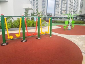 An outdoor exercise area with swings and a green exercise machine, surrounded by a red rubberized surface and greenery, with residential buildings in the background. - Clean And Nice 1Br At Apartment Casa De Parco (Tangerang)