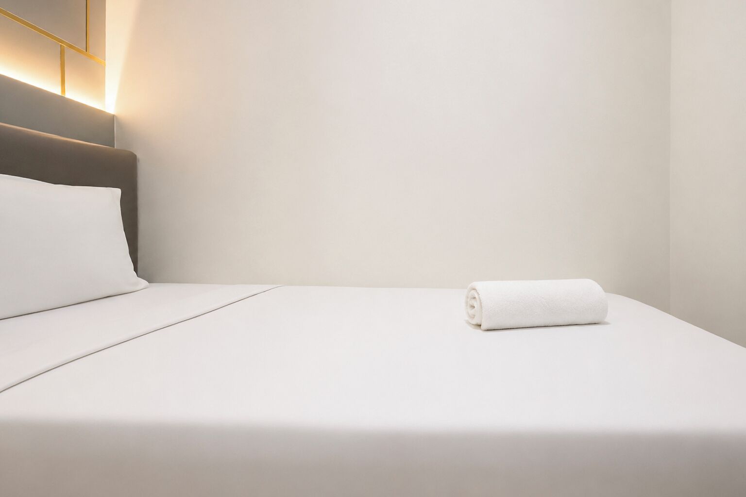 Een net opgemaakt bed met een wit kussen en een opgerolde witte handdoek, een witte, schone muur en een modern hoofdbord met een lamp.