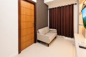A modern living room with a sofa, a wooden door, a curtain, and a television. - Functional With Minimalist Design 2Br At Vida View Makassar Apartment (Makassar)