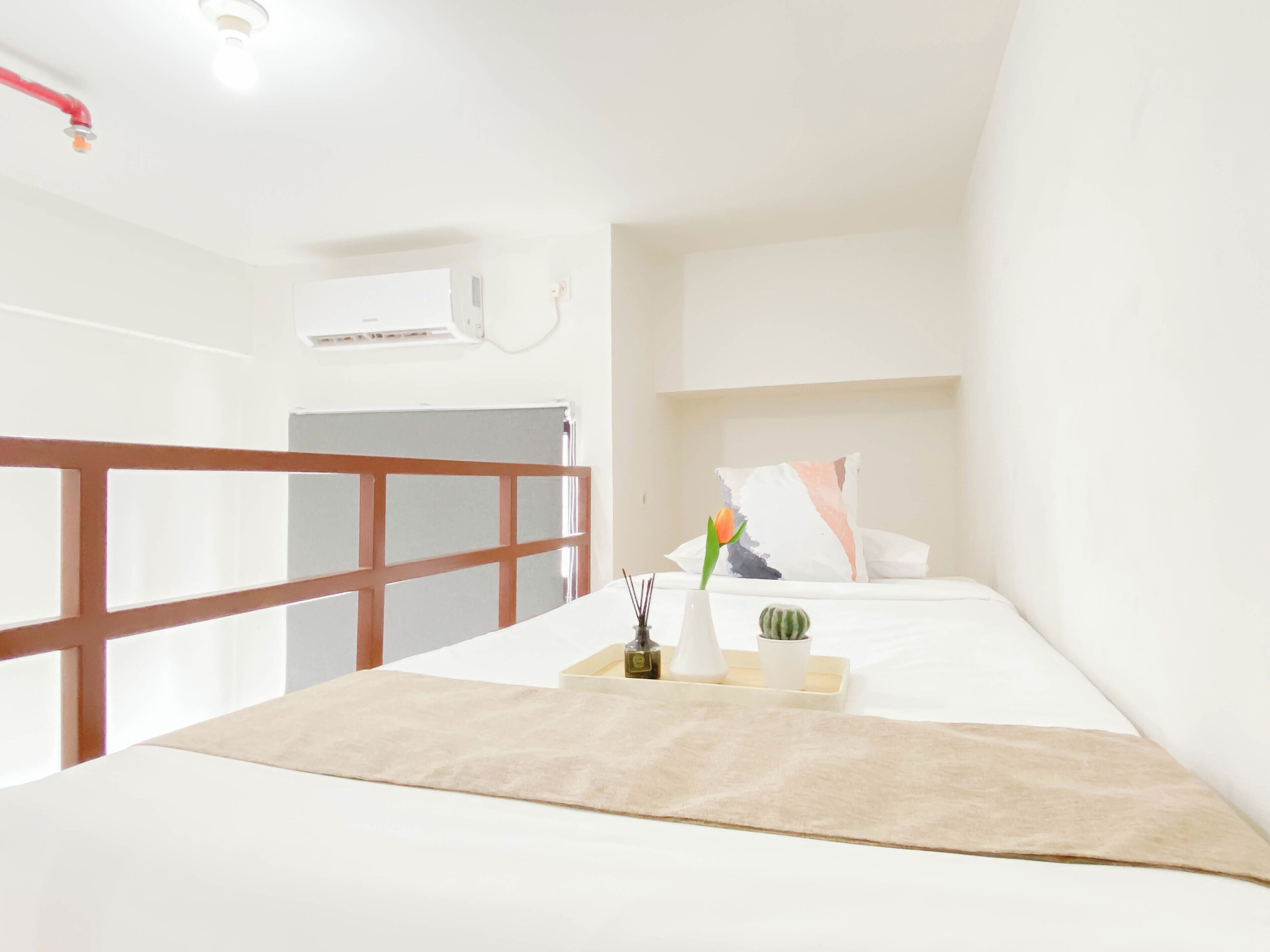 A hotel room with a bed, a bedside table with a vase and a plant, a window with blinds, and a white air conditioning unit on the wall.