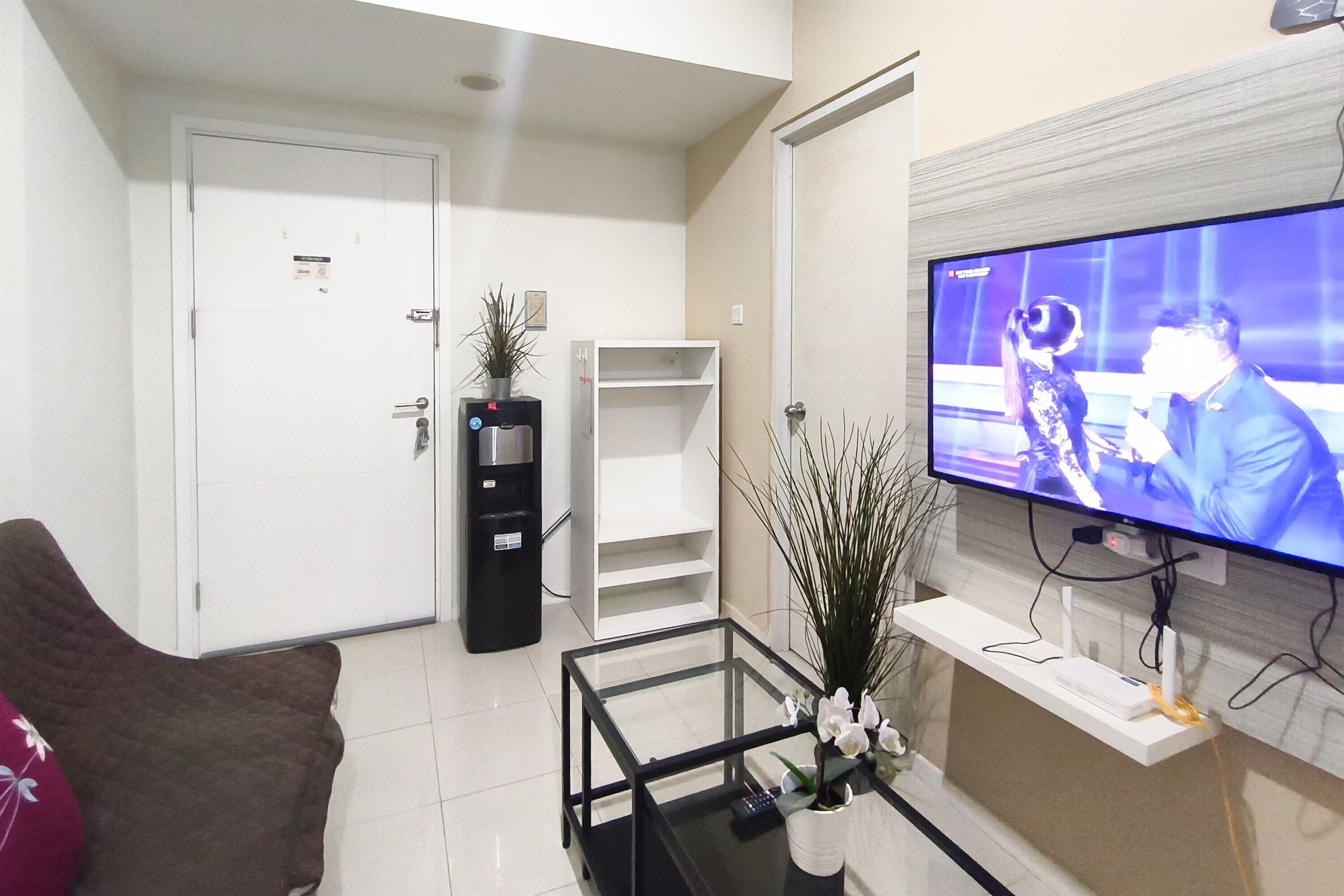 Una sala de estar con televisor de pantalla plana, una mesa de centro de vidrio, un sofá, una puerta blanca, un estante blanco y un mueble negro.