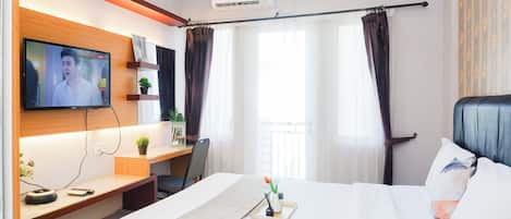 A hotel room with a bed, a desk with a TV, a plant, and a window with curtains.
