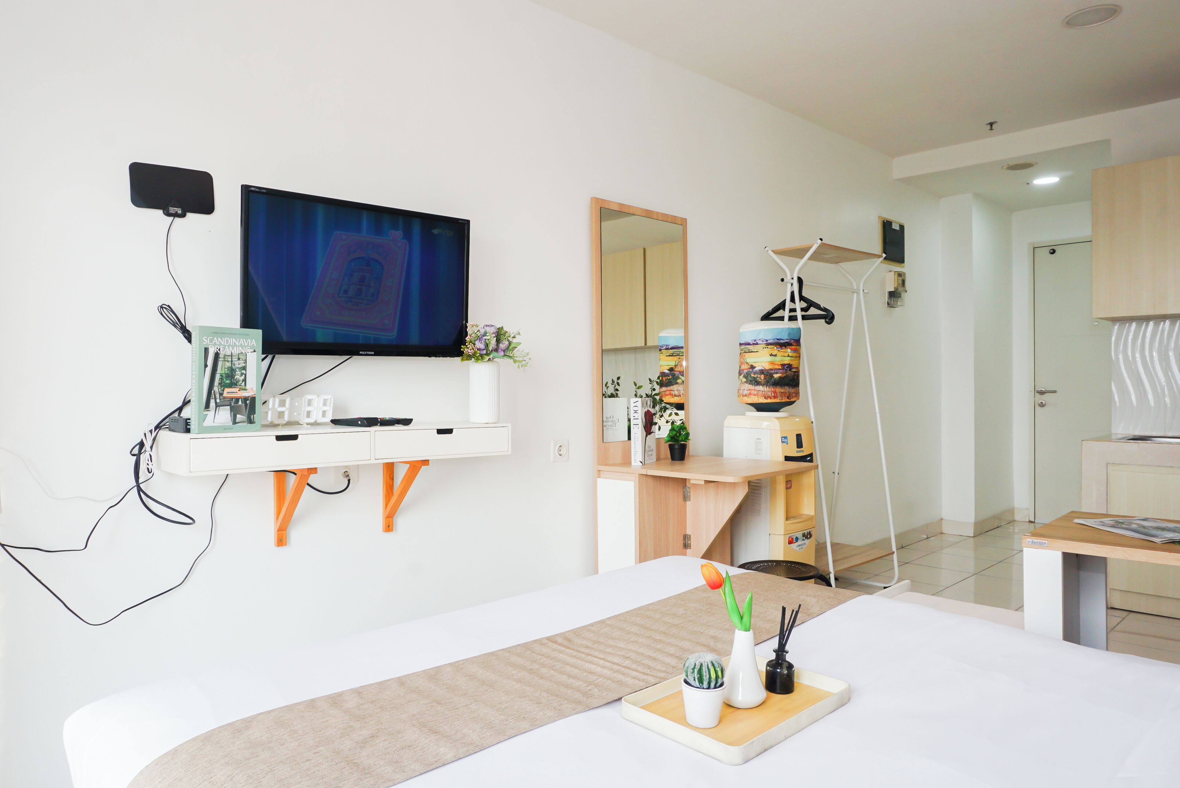 A modern hotel room with a flat-screen TV, a desk with a computer, a bed, a small table with a plant, and a mirror.