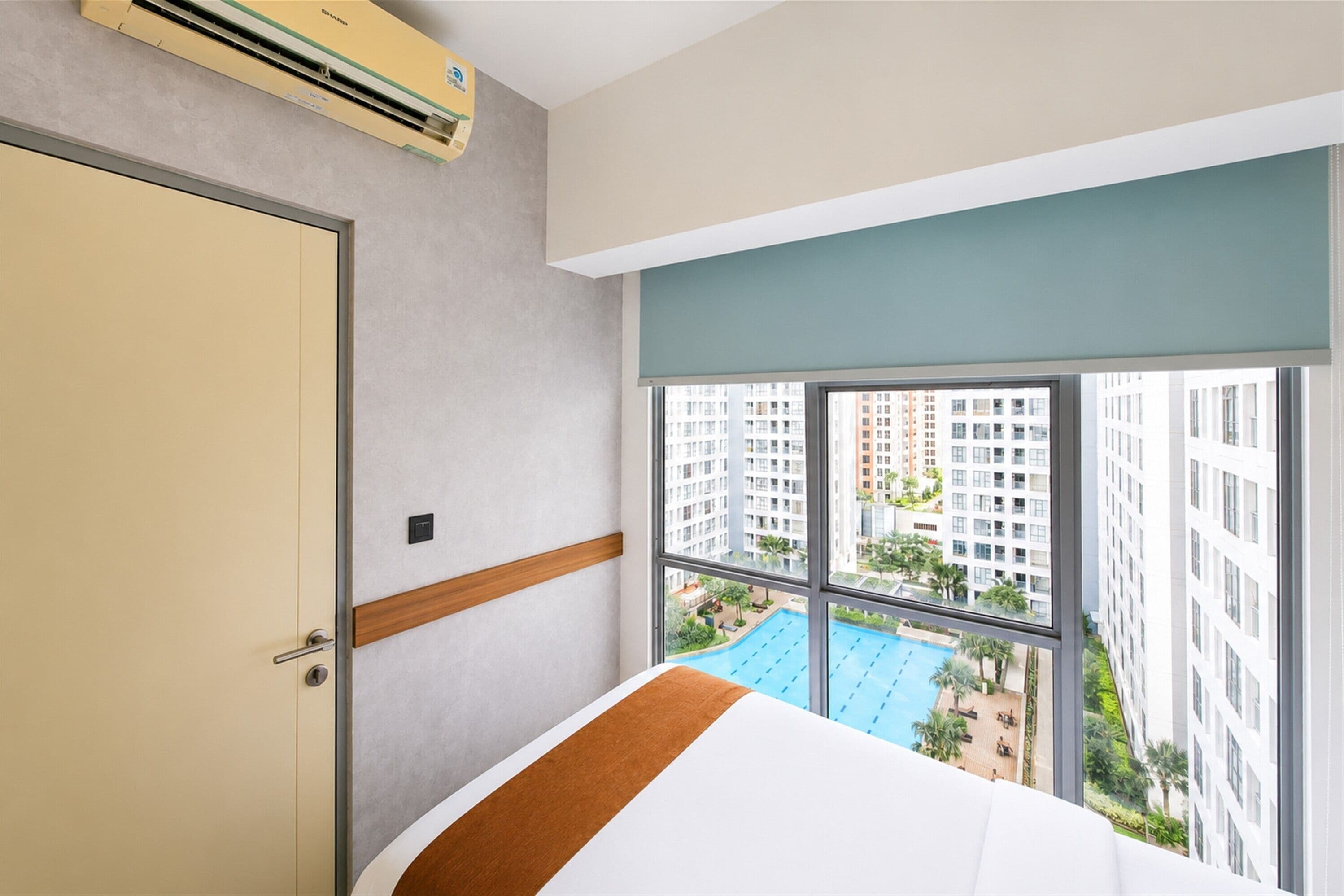 A hotel room with a large window, a bed, a wall-mounted air conditioner, and a view of a swimming pool and high-rise buildings.