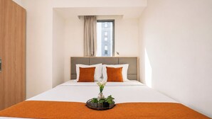 A hotel room with a bed, a window with curtains, a plant on the bed, and a wooden door. - Neat And Comfortable 1Br At Apartment M-Town Residence (Tangerang)