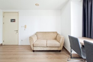 Ein Zimmer mit einem beigefarbenen Sofa, einem Holztisch, einem Stuhl, einer Tür mit einem Schild und einer Deckenleuchte.