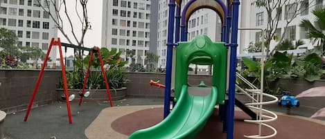 A playground with a green slide and red swings, surrounded by a paved area and greenery.