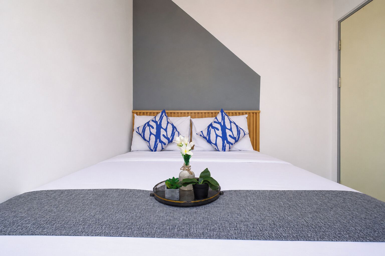 A neatly made bed with white bedding, a grey textured rug, and a tray with a vase of flowers.