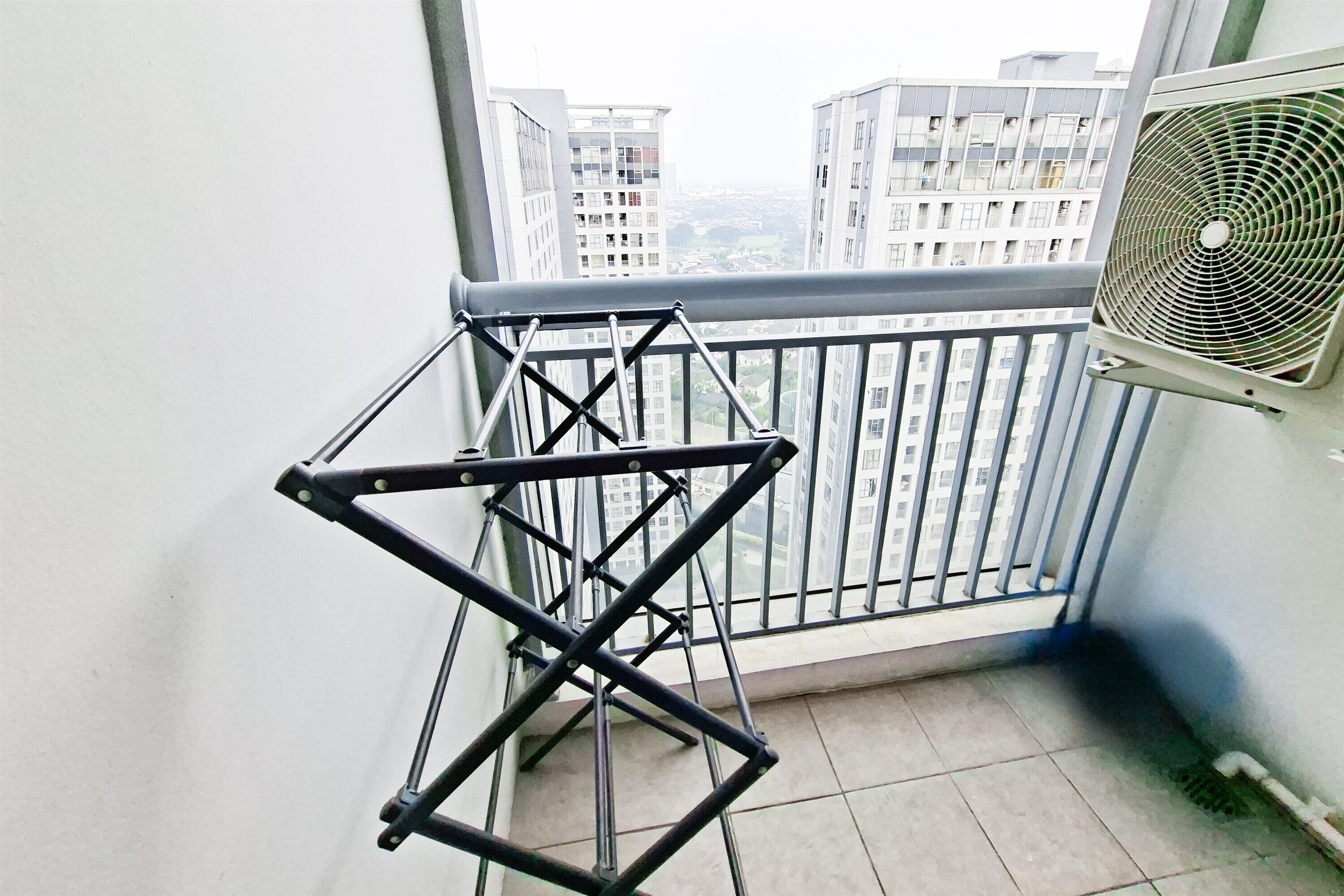 A balcony with a metal drying rack, a window with a view of high-rise buildings, and an air conditioning unit.